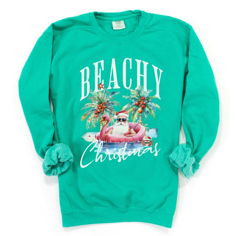 May include: Teal sweatshirt with a graphic of Santa Claus in sunglasses, floating on a pink flamingo in the ocean. Palm trees and the words "BEACHY Christmas" are above and below the illustration.