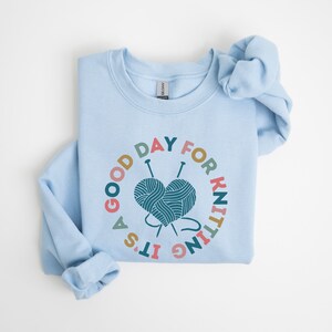 K&ouml;nnte beinhalten: Ein hellblaues Sweatshirt mit einer bunten Grafik, auf der "Es ist ein guter Tag zum Stricken" steht, mit einem herzf&ouml;rmigen Wollkn&auml;uel und Stricknadeln.