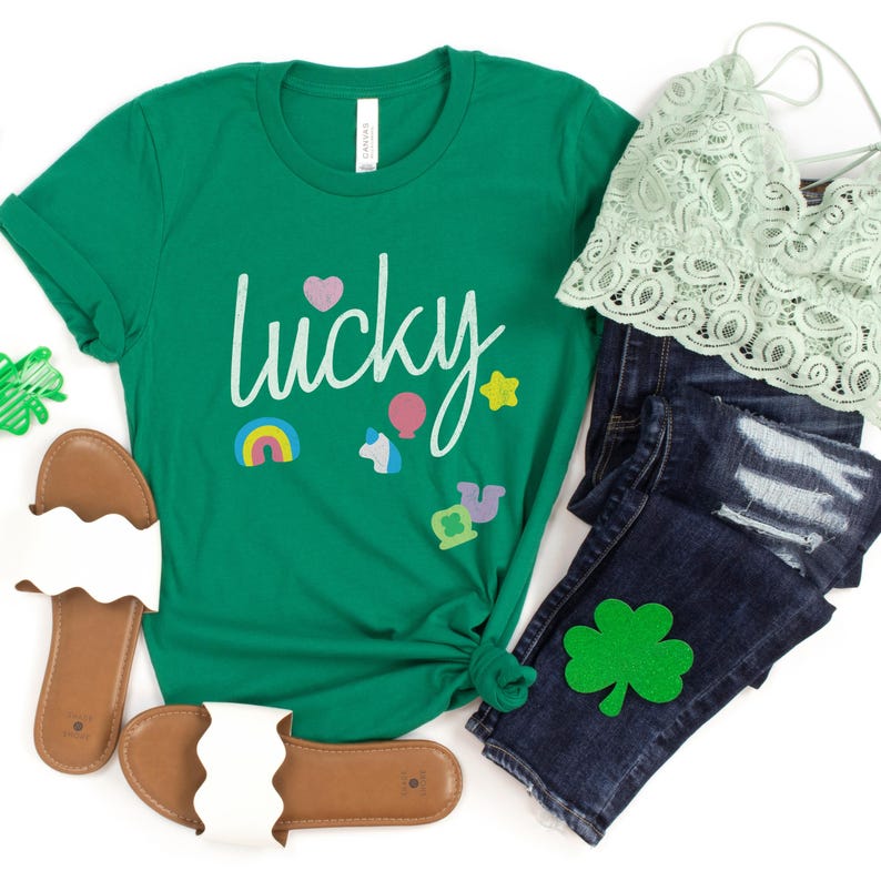 Puede incluir: Una camiseta verde con la palabra "lucky" escrita en blanco. La camiseta tiene un arco&iacute;ris, una estrella, una herradura, un coraz&oacute;n y un unicornio como gr&aacute;fico. La camiseta se combina con jeans azules, un parche de tr&eacute;bol verde y sandalias blancas.