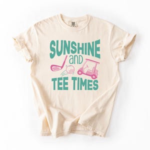 以下が含まれることがあります： クリーム色のTシャツ。 ターコイズとピンクで「SUNSHINE and TEE TIMES」の文字。 デザインにはゴルフクラブ、ゴルフボール、ゴルフカートが含まれています。 シャツは袖をまくり、クルーネックです。