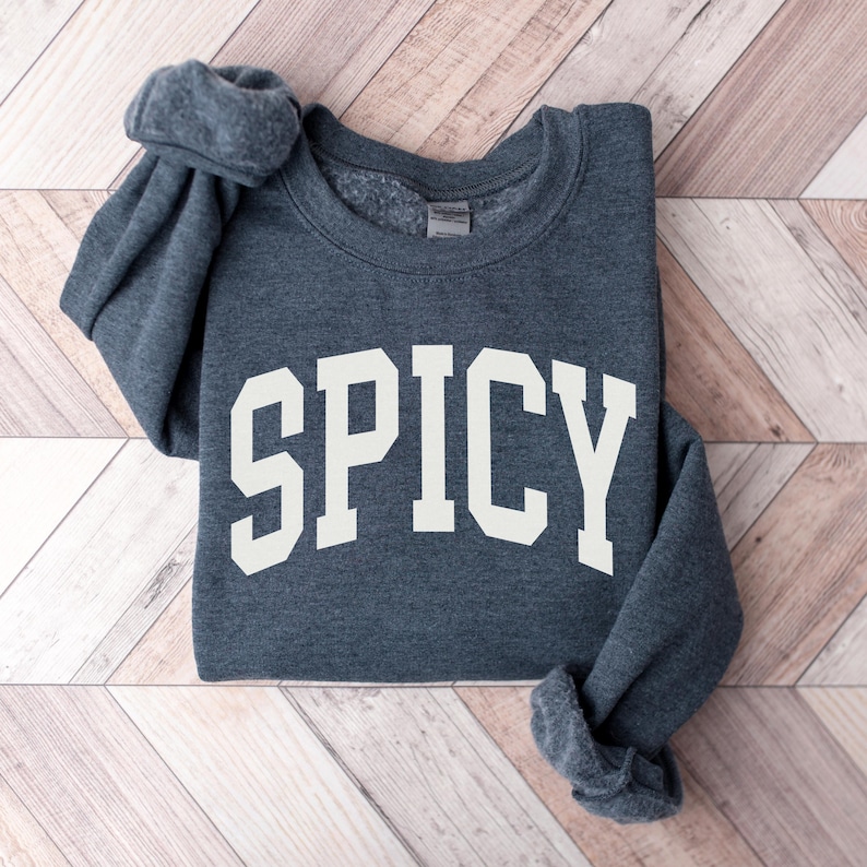 K&ouml;nnte beinhalten: Ein dunkelgrauer Pullover mit dem Wort "SPICY" in Wei&szlig; auf der Vorderseite.