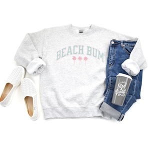 Pu&ograve; includere: Una felpa grigia con la scritta "Beach Bum" in turchese e palme rosa. La felpa &egrave; appoggiata su una superficie bianca con un paio di scarpe bianche da infilare, un paio di jeans blu e una tazza da viaggio grigia con la scritta "Rise & Grind" in bianco.