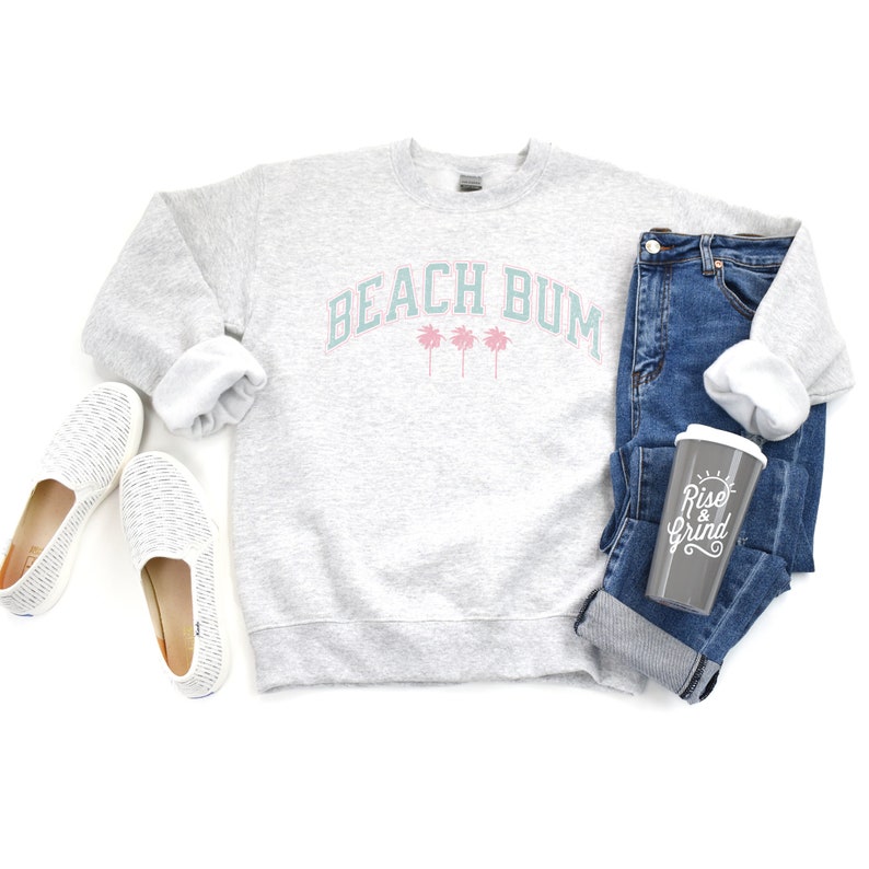 Pu&ograve; includere: Una felpa grigia con la scritta "Beach Bum" in turchese e palme rosa. La felpa &egrave; appoggiata su una superficie bianca con un paio di scarpe bianche da infilare, un paio di jeans blu e una tazza da viaggio grigia con la scritta "Rise & Grind" in bianco.