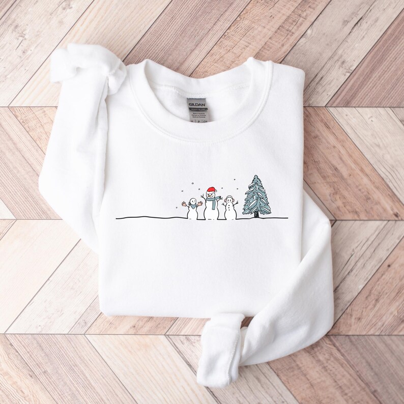 Op de afbeelding: Witte sweatshirt met een minimalistisch winterontwerp. De afbeelding toont drie sneeuwmannen en een kerstboom, allemaal in een eenvoudige lijntekeningstijl. E&eacute;n sneeuwman draagt een rode hoed en sjaal. Sneeuwvlokken zijn ook afgebeeld.