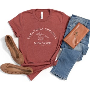 Puede incluir: Una camiseta de color marr&oacute;n rojizo con un dise&ntilde;o gr&aacute;fico blanco que dice "Saratoga Springs New York 1819" y presenta un jinete montando un caballo.
