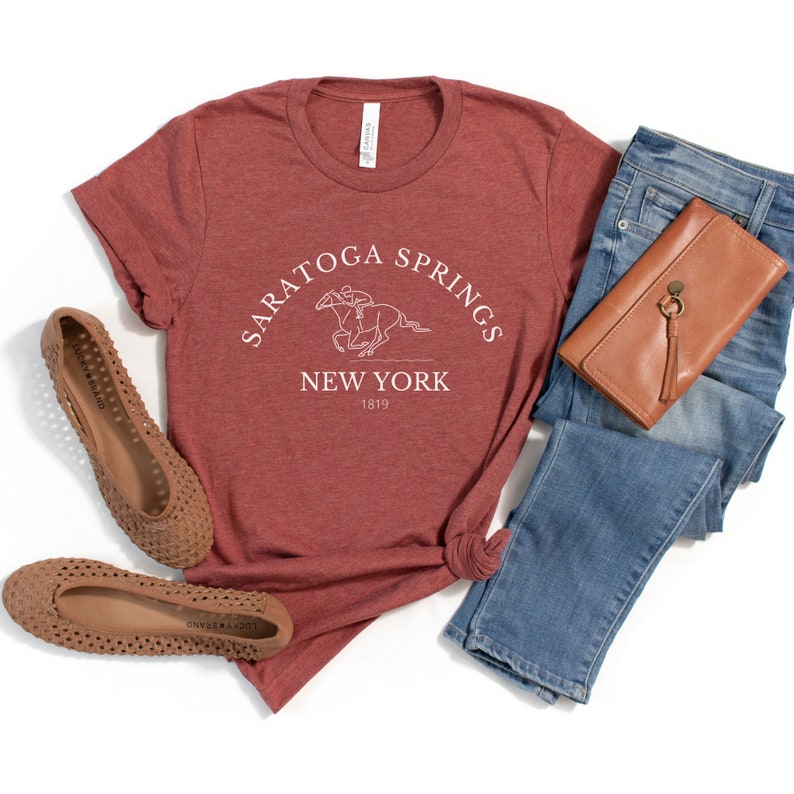 Puede incluir: Una camiseta de color marr&oacute;n rojizo con un dise&ntilde;o gr&aacute;fico blanco que dice "Saratoga Springs New York 1819" y presenta un jinete montando un caballo.