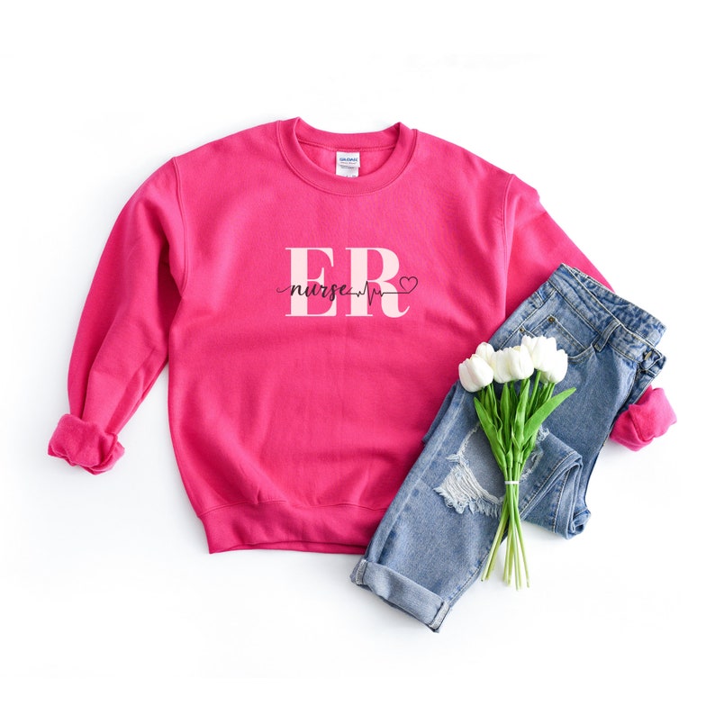 Puede incluir: Una sudadera rosa brillante con el texto "ER nurse" en fuente blanca, con una l&iacute;nea de latido card&iacute;aco y un s&iacute;mbolo de coraz&oacute;n. La sudadera se combina con vaqueros azules y un ramo de tulipanes blancos.