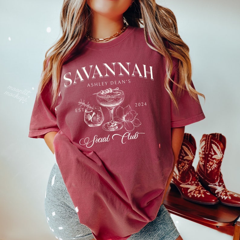 Pode incluir: Uma camiseta de cor vinho com texto branco que diz "Savannah Ashley Dean's Est. 2024 Social Club". O design inclui um copo de coquetel com uma guarni&ccedil;&atilde;o e uma flor.