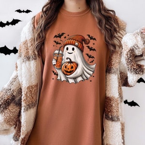 May include: A brown long-sleeved t-shirt with a cute ghost wearing a hat and holding a pumpkin and a cup of coffee. The ghost is surrounded by bats and stars.