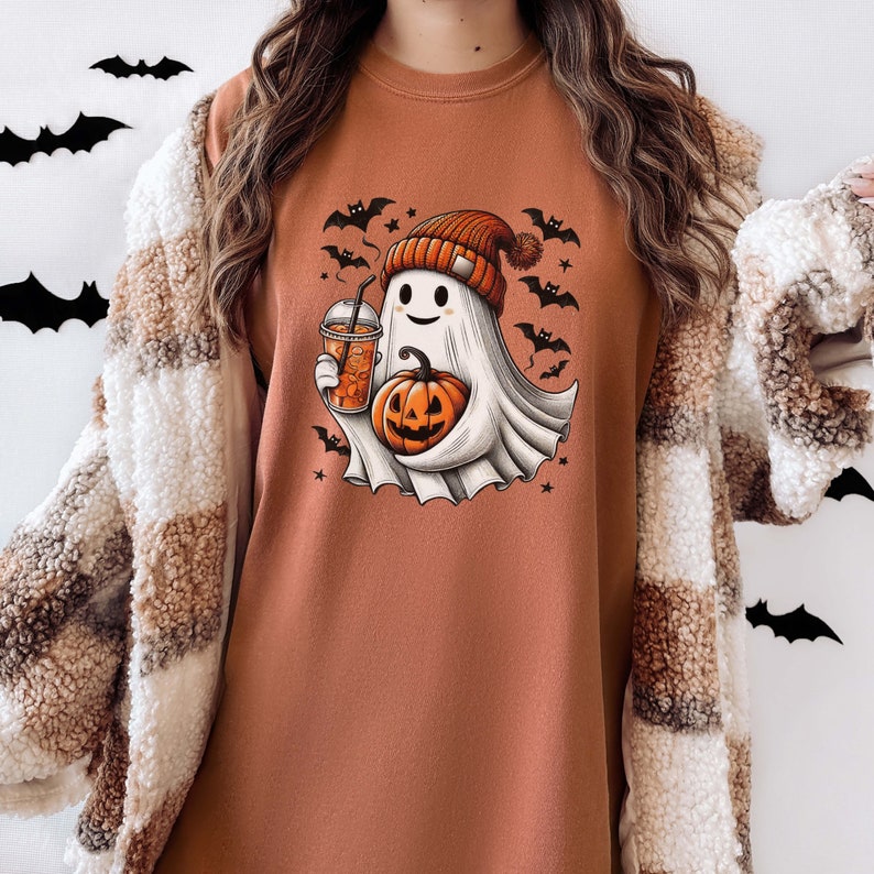 May include: A brown long-sleeved t-shirt with a cute ghost wearing a hat and holding a pumpkin and a cup of coffee. The ghost is surrounded by bats and stars.