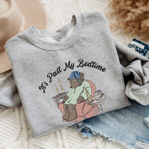 Pu&ograve; includere: Una felpa grigia con la scritta "It's Past My Bedtime" stampata su di essa. Un orso dei cartoni animati con un berretto da notte blu &egrave; seduto su una sedia con un libro e delle candele.