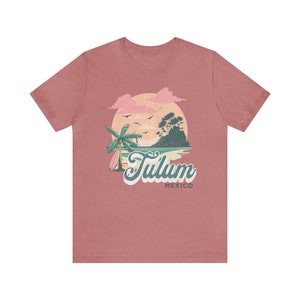 May include: A pink heather t-shirt with a graphic of a sunset over a beach with palm trees and the text "Tulum Mexico".