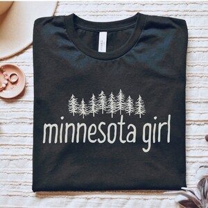 May include: Black t-shirt with a white graphic of a line of pine trees and the text "minnesota girl".