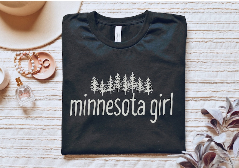 May include: Black t-shirt with a white graphic of a line of pine trees and the text "minnesota girl".