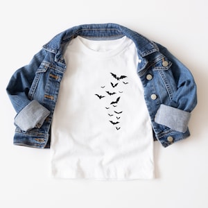 May include: White t-shirt with a black bat graphic. The bats are arranged in a descending diagonal pattern. A blue denim jacket is draped over the shirt.