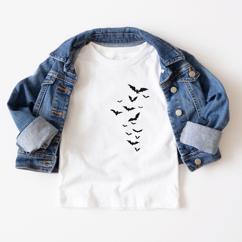May include: White t-shirt with a black bat graphic. The bats are arranged in a descending diagonal pattern. A blue denim jacket is draped over the shirt.