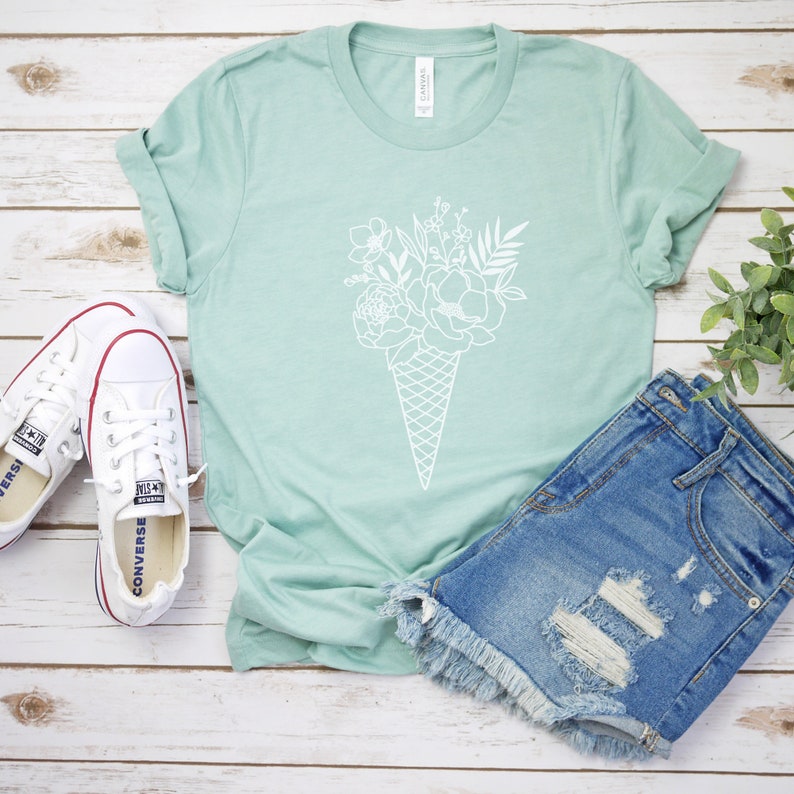 May include: A light green t-shirt featuring a white graphic of a bouquet of flowers in an ice cream cone. The t-shirt is styled with denim shorts and white trainers.