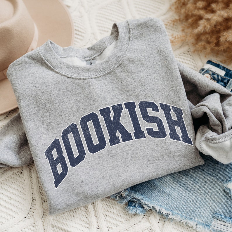 May include: Gray sweatshirt with the word "BOOKISH" printed in a navy blue, distressed, collegiate-style font.