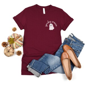 May include: A maroon t-shirt with a white ghost and the text "spooky season" printed on the front. The shirt is paired with blue jeans and brown flats.