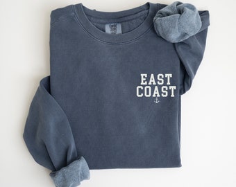 May include: A blue sweatshirt with the text "EAST COAST" in white lettering and an anchor symbol below. The sweatshirt is folded in half and the sleeves are rolled up.