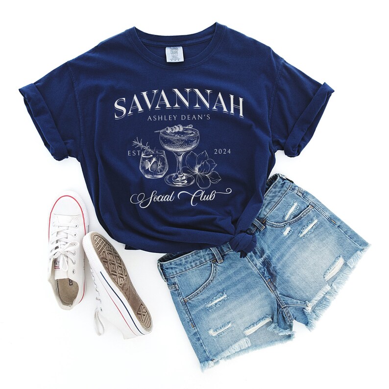 Pode incluir: Uma camiseta azul marinho com texto branco que diz "Savannah Ashley Dean's Est. 2024 Social Club". A camiseta apresenta um gr&aacute;fico de dois copos de coquetel com um design floral.
