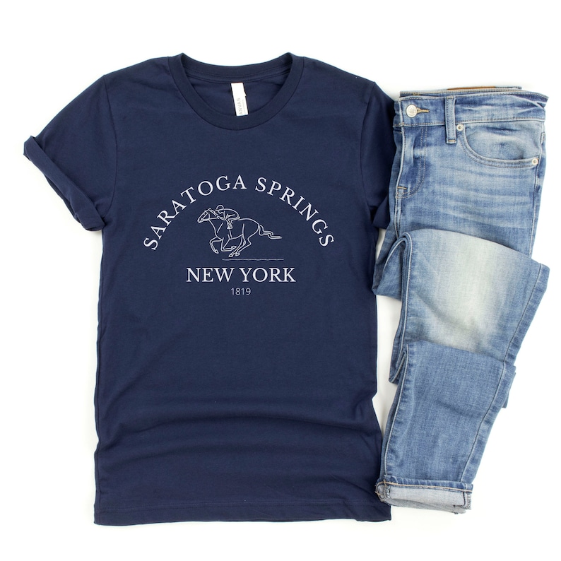 Puede incluir: Una camiseta azul marino con un gr&aacute;fico blanco de un caballo y un jinete. El gr&aacute;fico incluye el texto "Saratoga Springs New York 1819".