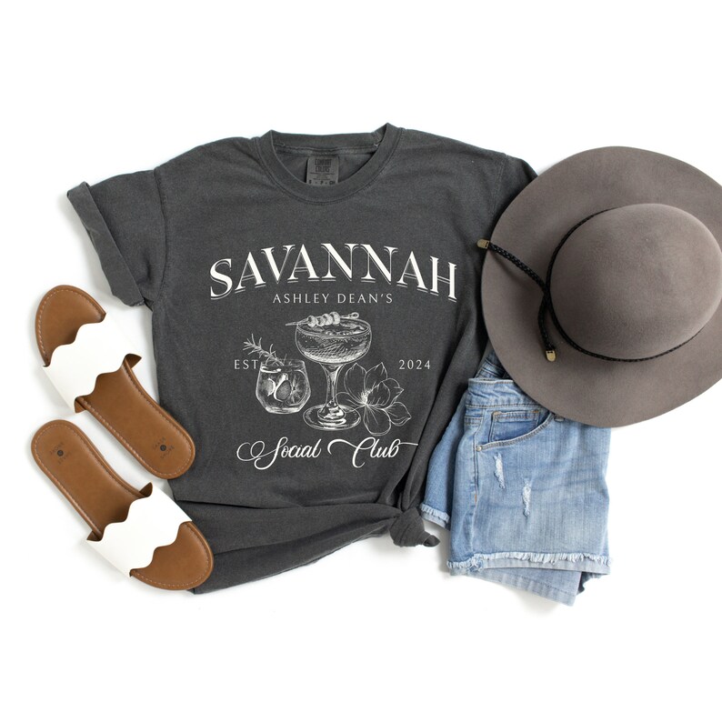Pode incluir: Uma camiseta cinza escura com um design gr&aacute;fico branco que diz "Savannah Ashley Dean's Est. 2024 Social Club". O gr&aacute;fico tamb&eacute;m inclui um copo de coquetel com uma bebida e uma flor.