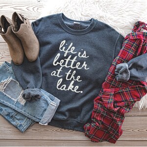 Op de afbeelding: Een donkergrijs sweatshirt met de tekst "life is better at the lake" in witte letters. Het sweatshirt ligt op een houten oppervlak met een paar bruine enkellaarsjes, een blauwe jeans en een rood-zwart geruit overhemd.