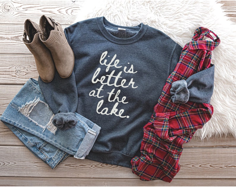 Op de afbeelding: Een donkergrijs sweatshirt met de tekst "life is better at the lake" in witte letters. Het sweatshirt ligt op een houten oppervlak met een paar bruine enkellaarsjes, een blauwe jeans en een rood-zwart geruit overhemd.