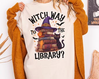 May include: Cream-colored t-shirt with the text "Witch Way to the Library?" featuring a witch's hat, stacked books, a jack-o'-lantern, and a black cat wearing glasses. Bats and stars are also included in the design.