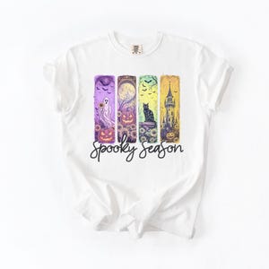 May include: White t-shirt featuring a colorful Halloween-themed design. The design includes a ghost, pumpkins, a black cat, and a spooky castle, with the words "Spooky Season" in a script font.