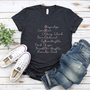 May include: A dark grey t-shirt with a white graphic design that spells out "BROOKLYN" using the names of different neighborhoods in Brooklyn, New York. The text is in a pink cursive font.