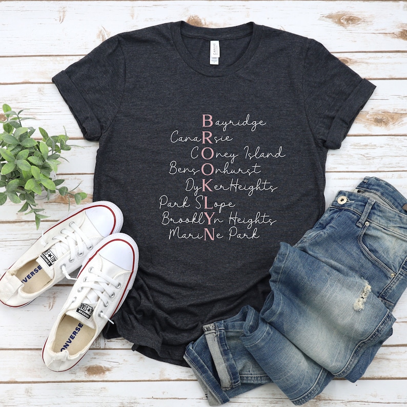 May include: A dark grey t-shirt with a white graphic design that spells out "BROOKLYN" using the names of different neighborhoods in Brooklyn, New York. The text is in a pink cursive font.