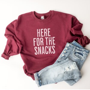 May include: A maroon sweatshirt with the text "HERE FOR THE SNACKS" in white lettering. The sweatshirt is paired with light blue jeans.