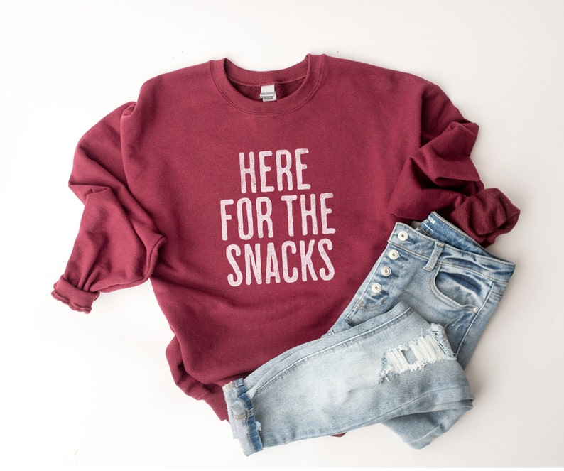 May include: A maroon sweatshirt with the text "HERE FOR THE SNACKS" in white lettering. The sweatshirt is paired with light blue jeans.