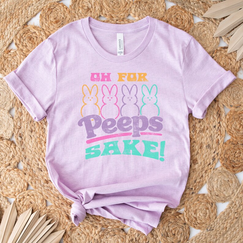 May include: A lavender t-shirt with the text "Oh for Peeps Sake!" in a retro font. Four colorful Peeps marshmallow chicks are arranged in a row above the text.