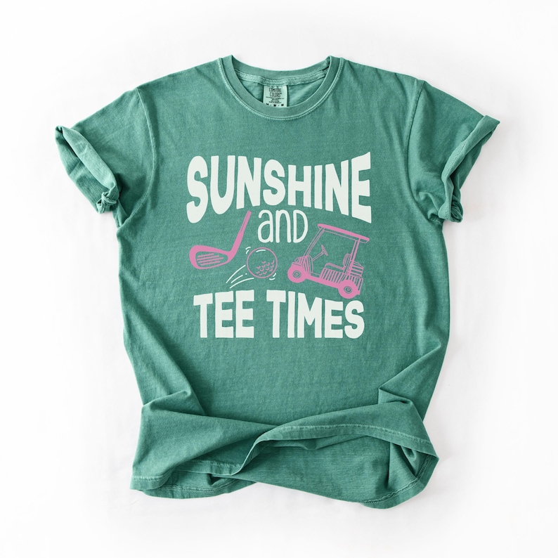 以下が含まれることがあります： 「SUNSHINE and TEE TIMES」の白い文字が入った、ティールグリーンのTシャツ。ゴルフのクラブ、ゴルフボール、ゴルフカートのピンク色のグラフィックも特徴です。このシャツは柔らかい素材で作られています。