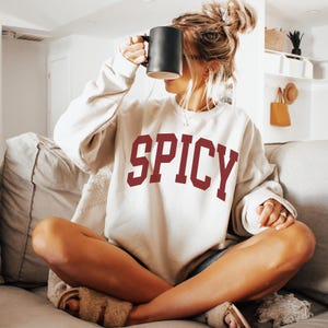 Może przedstawiać: Kobieta w białej bluzie z nadrukiem "SPICY" w kolorze bordowym na przodzie. Siedzi na kanapie i trzyma czarny kubek.