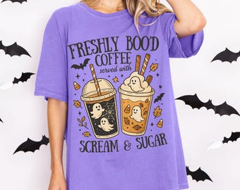 May include: A purple t-shirt with the text "FRESHLY BOO'D COFFEE served with SCREAM & SUGAR." The graphic features two coffee cups with ghost designs, leaves, and bats. The shirt is a comfortable, oversized fit.