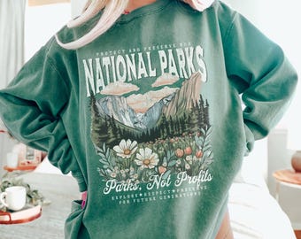 Könnte beinhalten: Grünes Sweatshirt mit dem Text "PROTECT AND PRESERVE OUR NATIONAL PARKS". Die Grafik zeigt eine Berglandschaft mit Blumen und dem Text "Parks, Not Profits. Explore, Respect, Preserve for Future Generations."
