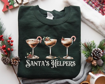 Può includere: Felpa verde scuro con tre bicchieri da cocktail con bevande color caffè, bastoncini di zucchero e agrifoglio. Il testo "Santa's Helpers" è stampato sotto. Abbigliamento festivo per le vacanze.