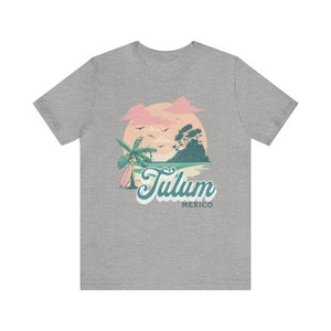 May include: A heather grey t-shirt with a graphic of a sunset over a beach with palm trees and a surfboard. The text "Tulum Mexico" is printed below the graphic.