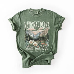 K&ouml;nnte beinhalten: Gr&uuml;nes T-Shirt mit einem Vintage-Grafikdruck einer Berglandschaft und Wildblumen. Der Text lautet "Sch&uuml;tzen und bewahren Sie unsere Nationalparks" und "Parks, nicht Profite. Erkunden. Respektieren. Widerstehen."