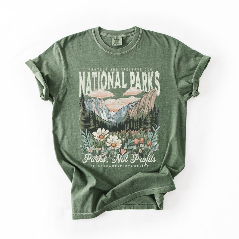 K&ouml;nnte beinhalten: Gr&uuml;nes T-Shirt mit einem Vintage-Grafikdruck einer Berglandschaft und Wildblumen. Der Text lautet "Sch&uuml;tzen und bewahren Sie unsere Nationalparks" und "Parks, nicht Profite. Erkunden. Respektieren. Widerstehen."