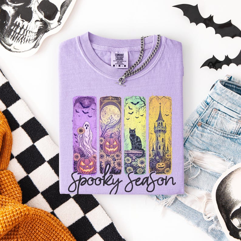May include: A lavender Comfort Colors t-shirt with a silver chain necklace. The shirt features a "Spooky Season" graphic with a ghost, pumpkin, black cat, and a castle. The shirt is folded on a white surface with Halloween decorations.