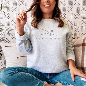 Op de afbeelding: Een grijs sweatshirt met een eenvoudige lijntekening van een halve maan, sterren en een planeet. De tekst "TRUST THE UNIVERSE" is onder de tekening gedrukt.