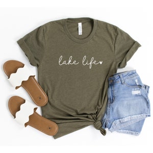 K&ouml;nnte beinhalten: Ein gr&uuml;nes T-Shirt mit dem Text "lake life" in wei&szlig;en Buchstaben und einem kleinen wei&szlig;en Herz. Das Shirt liegt flach auf einem wei&szlig;en Hintergrund mit einem Paar wei&szlig;en Sandalen und einer Jeans-Shorts.