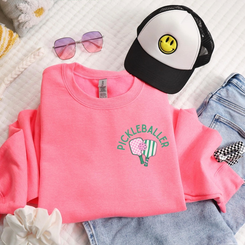 May include: Pink sweatshirt with a green and pink pickleball paddle graphic and the text "Pickleballer" on the front.