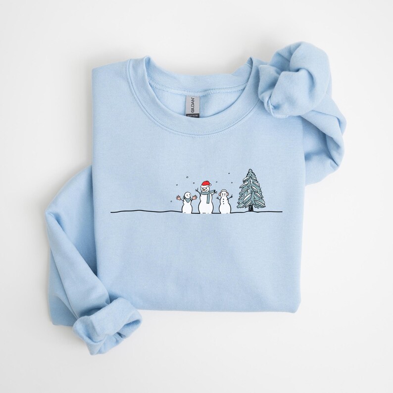 Op de afbeelding: Lichtblauw sweatshirt met een grillige wintersc&egrave;ne. Het ontwerp toont drie sneeuwmannen, een kerstboom en sneeuwvlokken. De middelste sneeuwman draagt een rode hoed en sjaal. De sc&egrave;ne is getekend op een zwarte lijn.