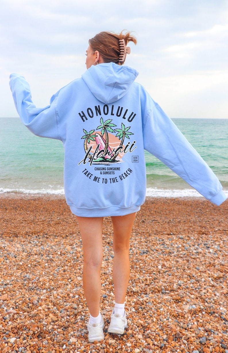 May include: A light blue hoodie with a graphic print on the back. The graphic features palm trees, a surfboard, and the text "Honolulu Hawaii" with the words "Chasing Sunshine & Sunsets Take Me To The Beach" below.